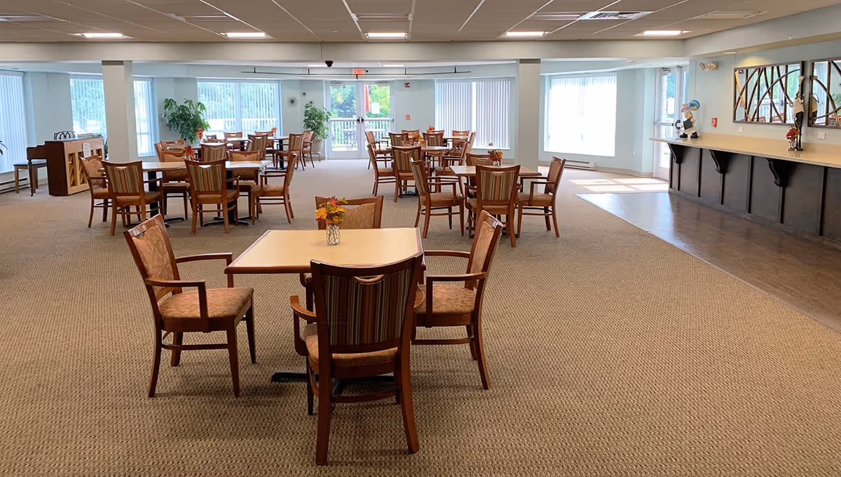 Spacious dining/activity room with multiple tables and chairs, a service counter on the right, and large windows providing natural light.