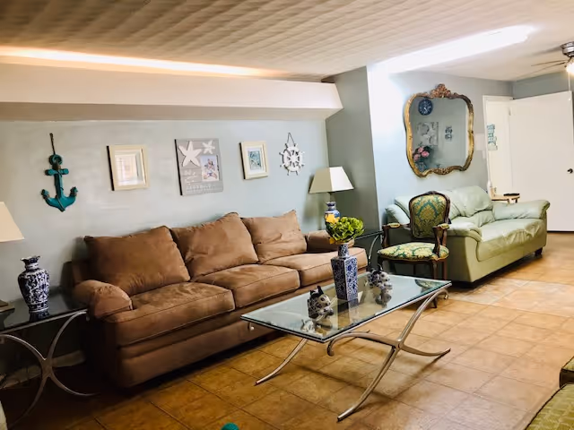 A cozy living room with a brown sofa, a green armchair, and a light green loveseat. The room features a glass coffee table with decorative items on it, a side table with a blue and white vase, and wall decorations including framed pictures, a green anchor, and a large ornate mirror. The floor is tiled and the walls are painted light blue.