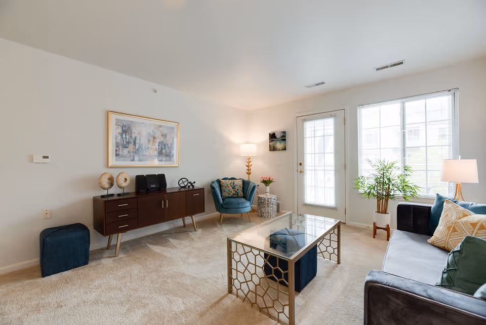 A bright and cozy living room with beige carpet, a gray sofa with colorful pillows, a glass coffee table with a gold geometric frame, a blue armchair, a wooden sideboard with decorative items, a floor lamp, and large windows letting in natural light.