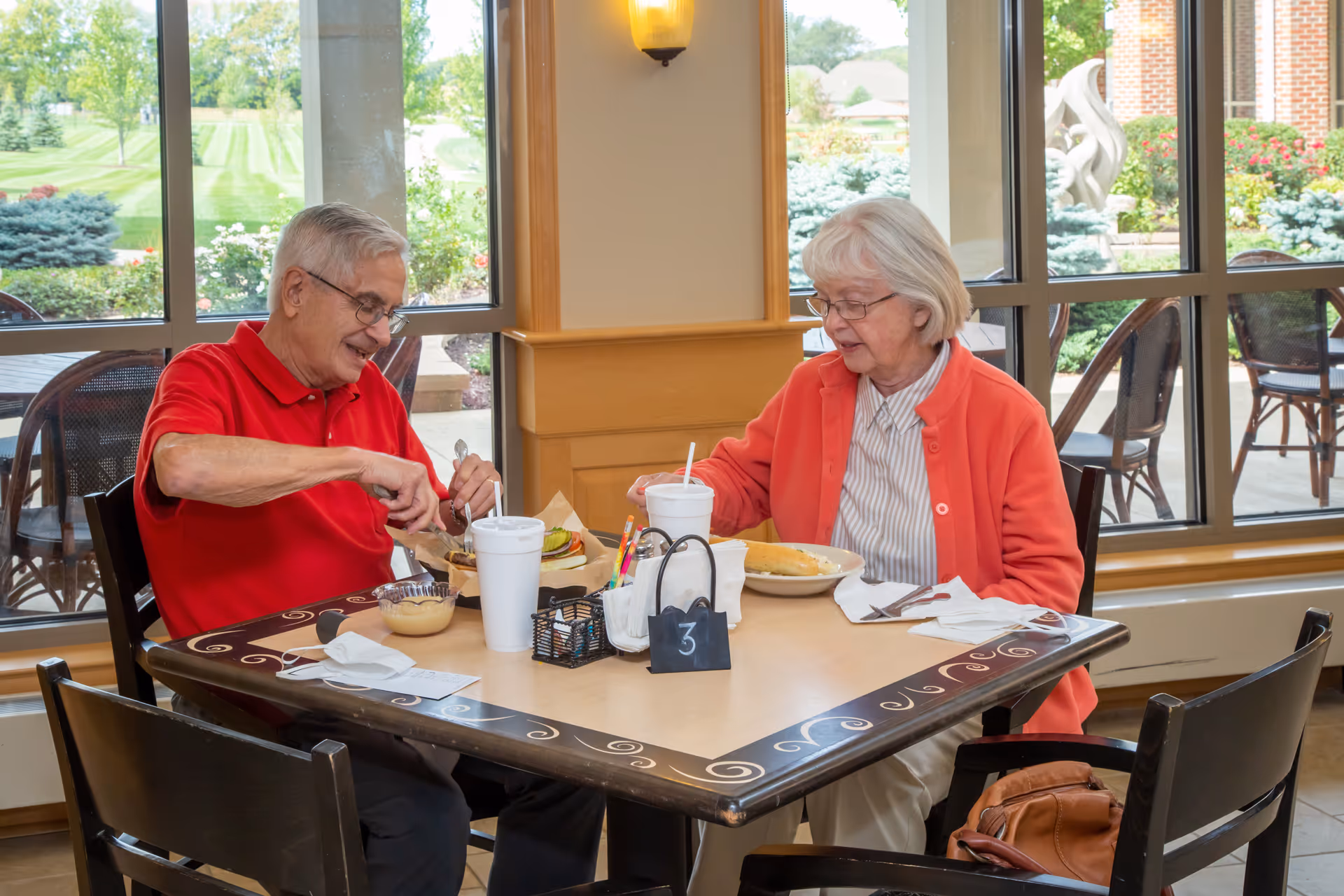 An elderly man and woman sitting at a dining table inside a facility, enjoying a meal together. The man is wearing a red polo shirt and glasses, cutting his food, while the woman, also wearing glasses and a red cardigan, is looking at her plate. The table has drinks, utensils, napkins, and a small table number sign. Large windows behind them show a green outdoor garden area.