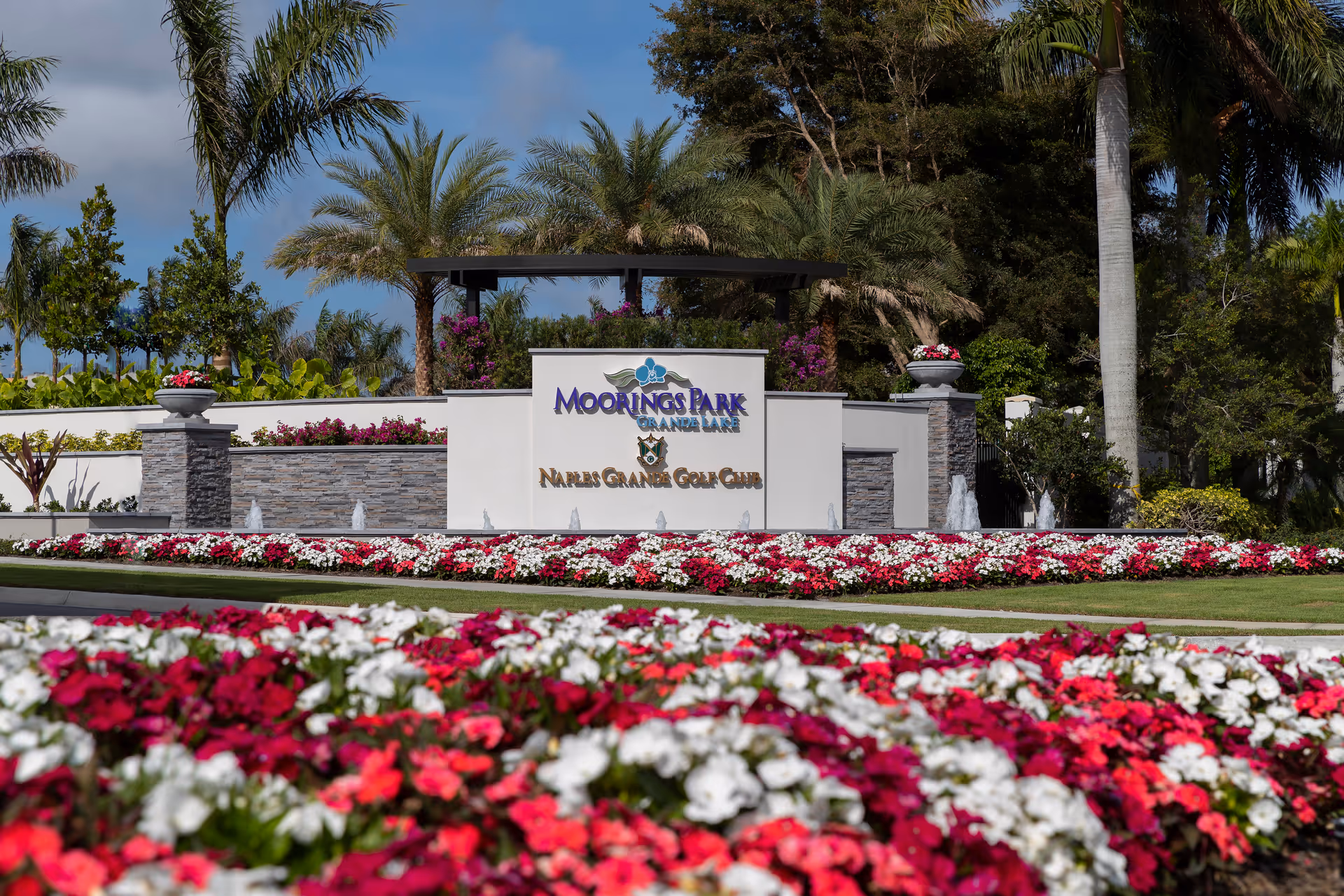 Entrance sign for Moorings Park Grande Lake and Naples Grande Golf Club surrounded by colorful flower beds and palm trees under a blue sky.