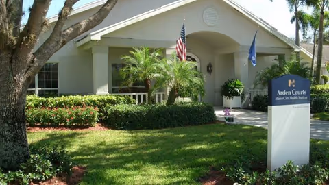 Exterior view of Arden Courts - ProMedica Memory Care Community in Delray Beach, showing a single-story building with a well-maintained lawn, palm trees, and two flags near the entrance. A sign with the facility's name is visible in the foreground.