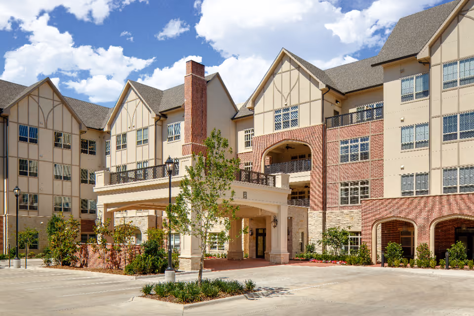 Exterior view of a multi-story assisted living facility with a covered entrance, brick and beige facade, multiple windows, and landscaped greenery under a partly cloudy sky.