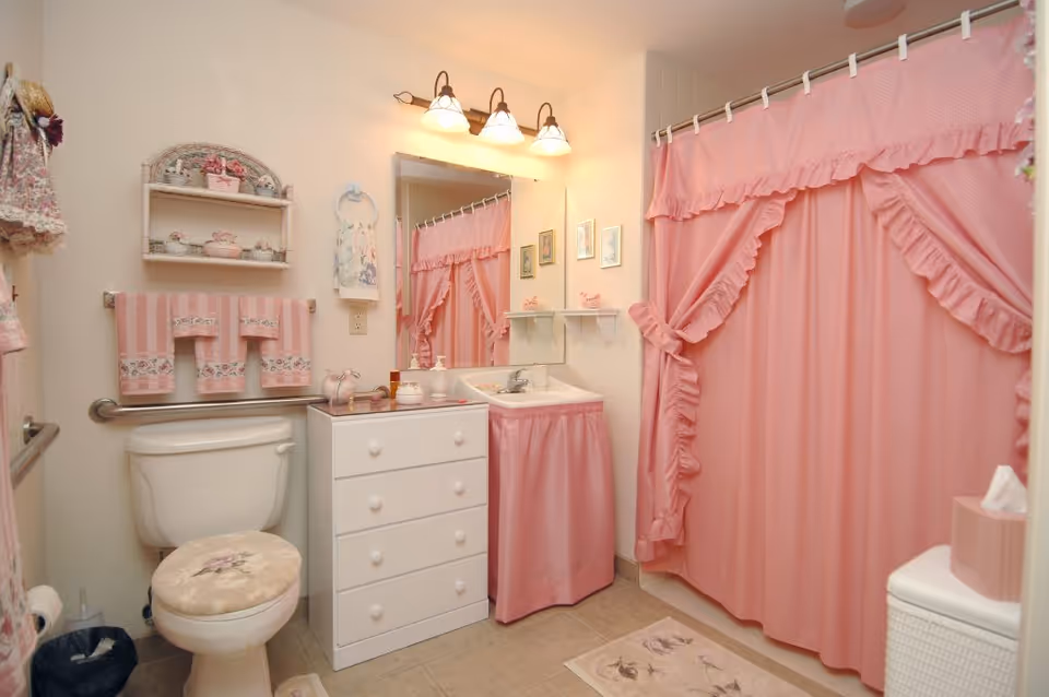 A bathroom with a white toilet, a white vanity with drawers, and a sink covered with a pink ruffled skirt. The shower has a pink ruffled curtain, and there are pink towels hanging on the wall. The bathroom is decorated with floral and pink accents, including a cushioned toilet seat cover, a small shelf with decorative items, and a pink tissue box on a white wicker cabinet.