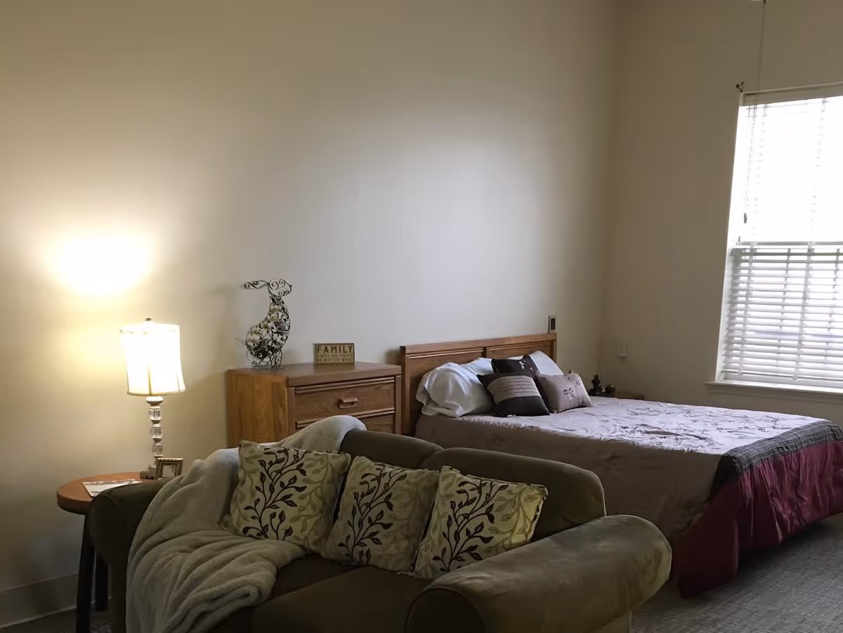 A cozy bedroom with a bed made up with pillows and a quilt, a wooden dresser with decorative items on top, a green couch with patterned cushions and a blanket, a side table with a lamp, and a window with blinds letting in natural light.