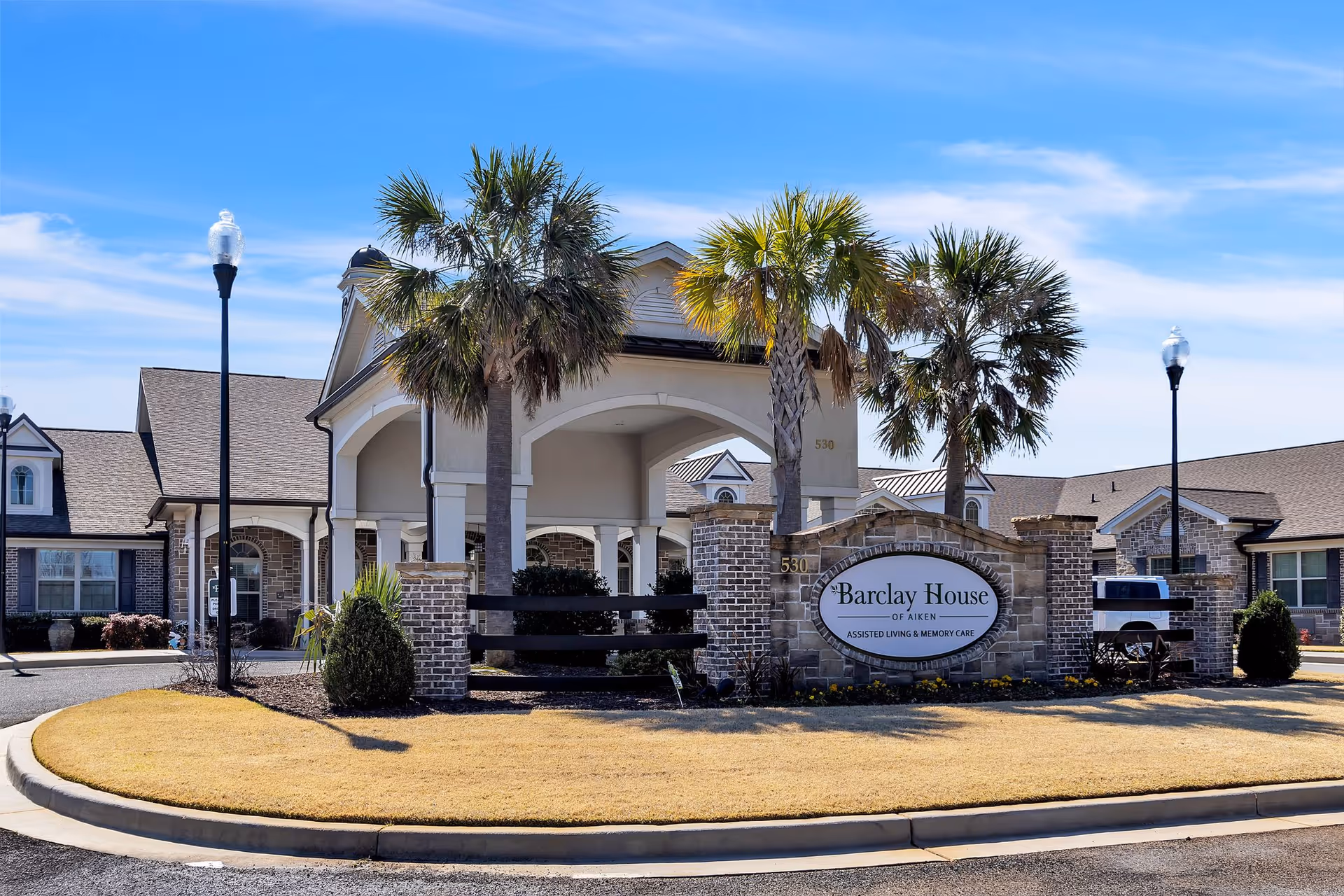 Exterior view of Barclay House of Aiken, an assisted living and memory care facility, showing the entrance with a covered driveway, palm trees, a brick sign with the facility name, and surrounding buildings under a clear blue sky.