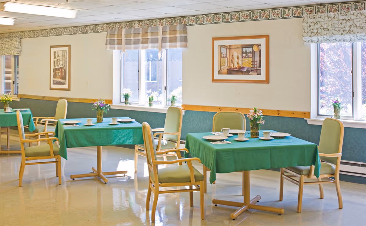 A bright dining room with several tables covered in green tablecloths, each set with white cups, plates, and utensils. The room has large windows with floral and plaid valances, framed artwork on the walls, and wooden chairs with green cushions around the tables. Small flower arrangements are placed on each table and windowsill.