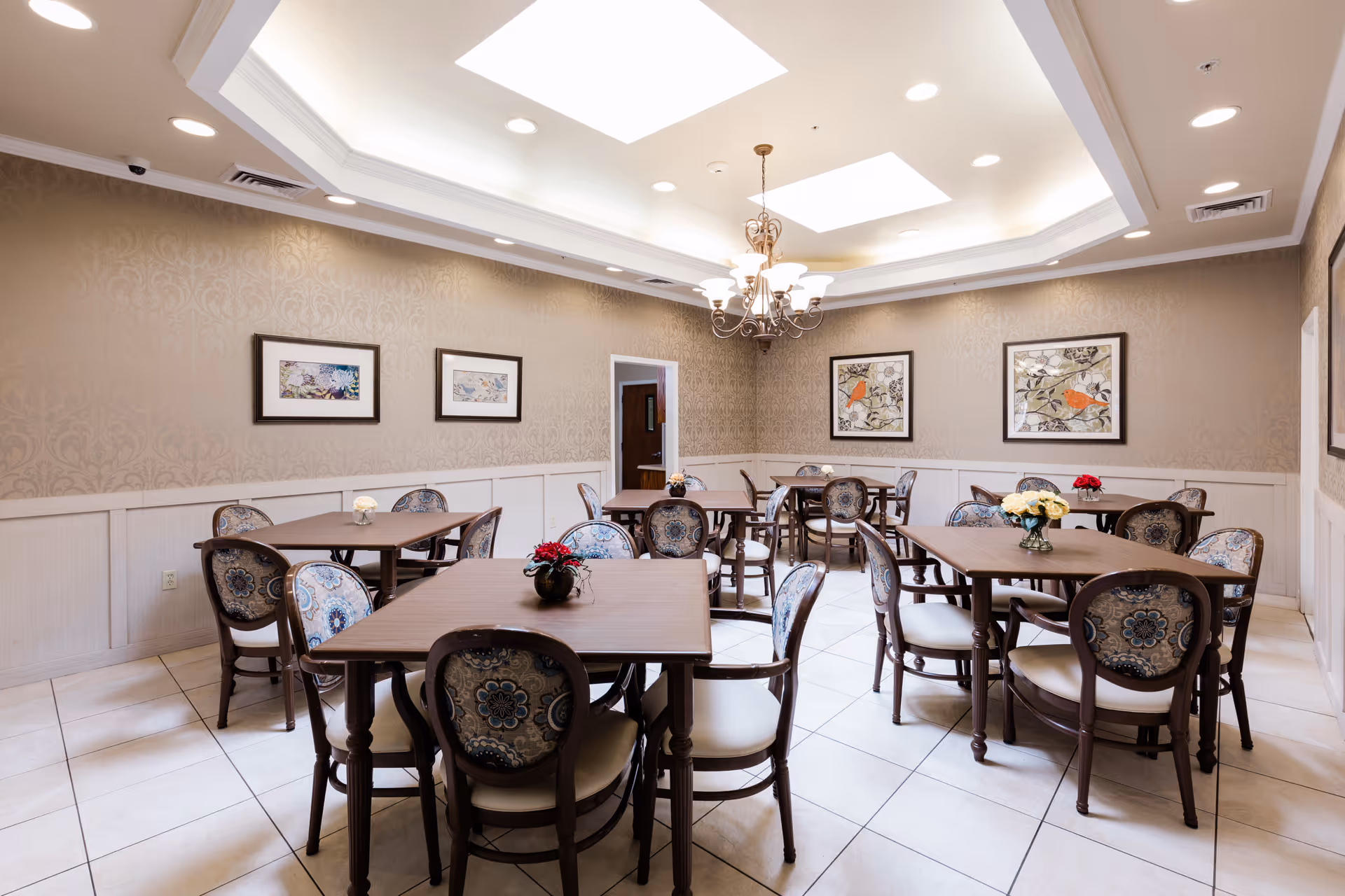 A well-lit dining room with several square wooden tables and cushioned chairs featuring floral upholstery. The walls have a beige patterned wallpaper with white wainscoting, and framed artwork is hung on the walls. A chandelier hangs from the ceiling with recessed lighting and skylights providing additional light. Small flower arrangements are placed on each table.