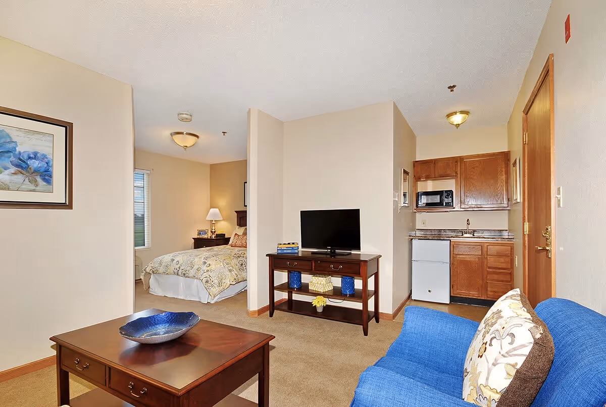 Interior view of a senior living facility room at Virginia Place showing a living area with a blue armchair and wooden coffee table, a TV on a wooden stand, a small kitchenette with a mini fridge, microwave, and cabinets, and a partially visible bedroom with a bed, nightstand, and lamp.