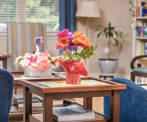 Cozy seating area with a wooden side table displaying a pink-wrapped pot of orange and pink flowers, magazines on the lower shelf, and upholstered chairs and plants in the background.