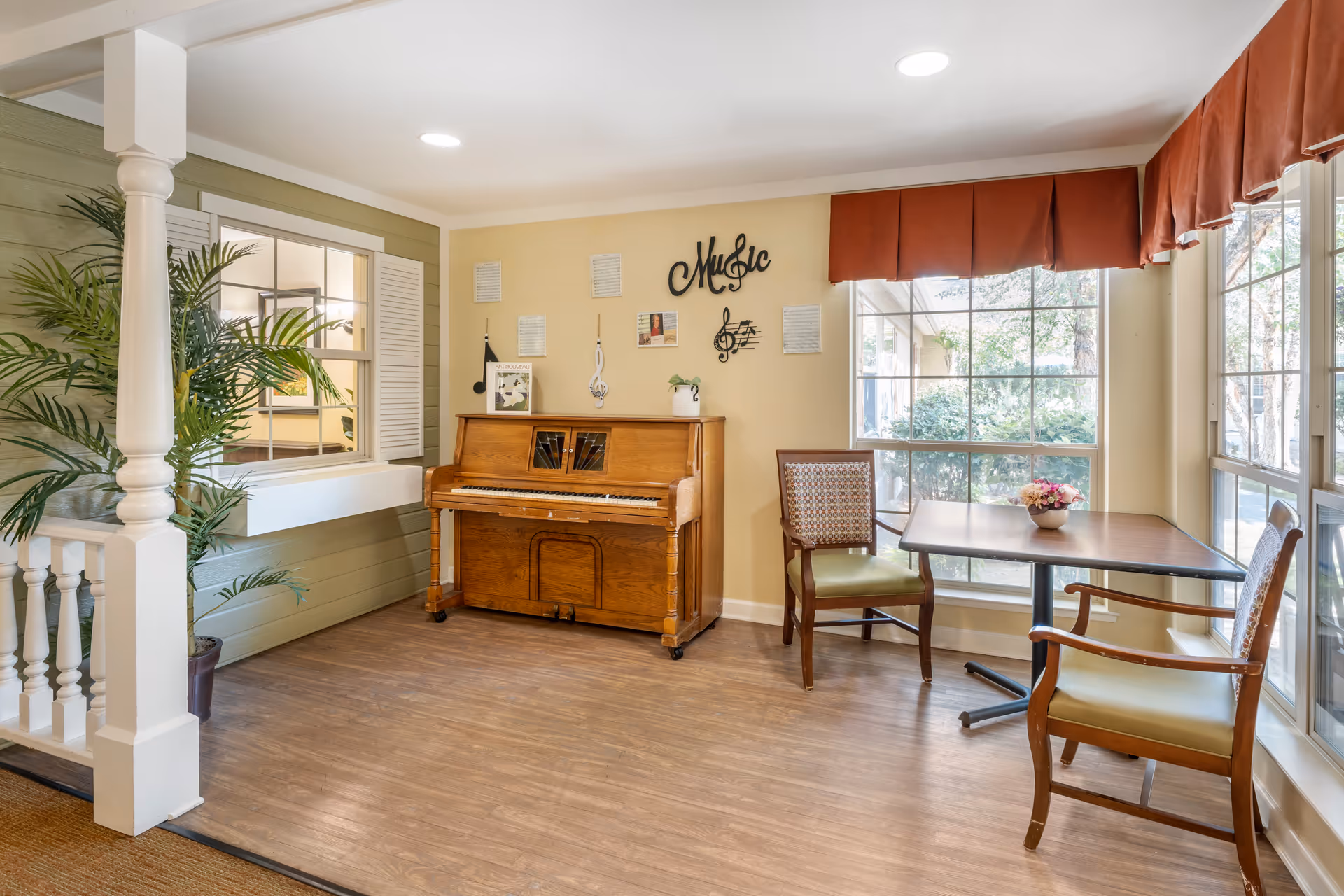 A cozy interior corner of a senior living facility featuring a wooden upright piano against a beige wall decorated with musical notes and the word 'Music'. Next to the piano are two wooden chairs with green cushions and a small table with a floral arrangement. Large windows with red valances let in natural light, and a potted plant is placed near a white column and railing.