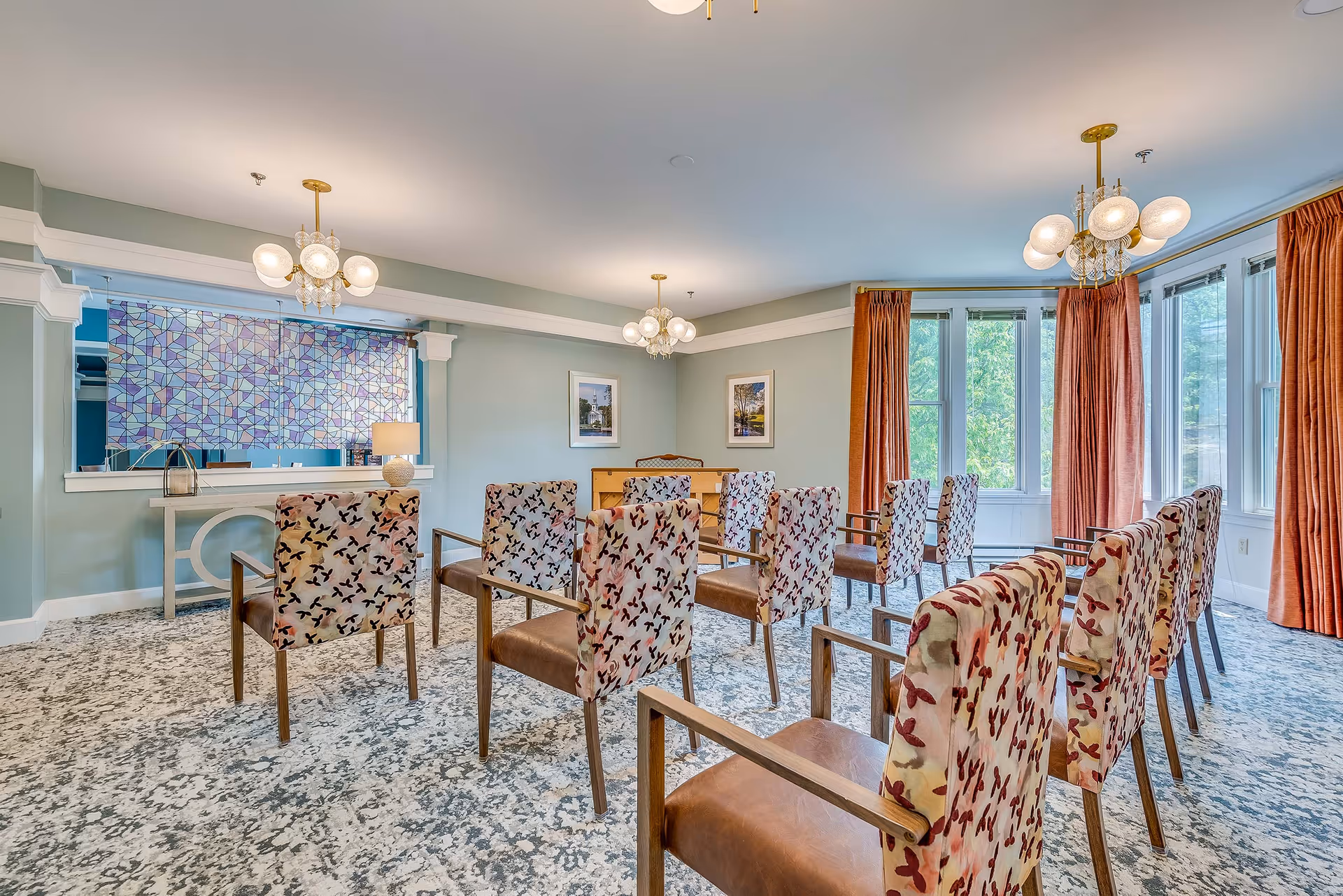A bright room with multiple patterned chairs arranged in rows facing a piano against a light blue wall. The room features large windows with orange curtains, three modern chandeliers, a patterned carpet, and framed pictures on the walls. A decorative console table with a lamp and a colorful stained glass window panel are visible on the left side.