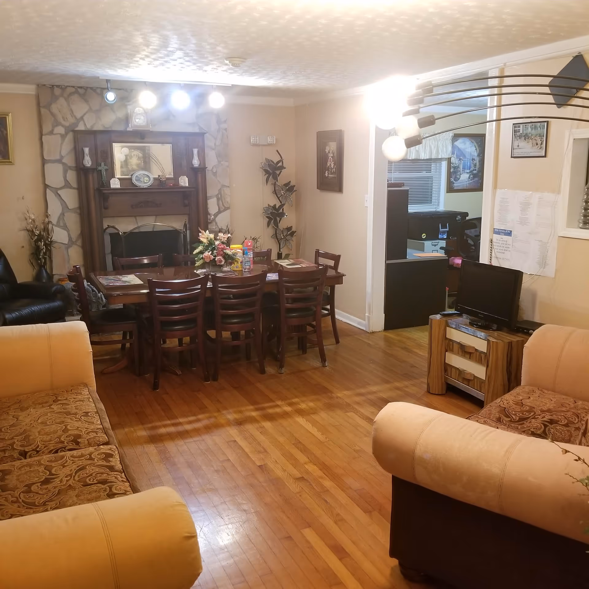 A cozy living and dining area with wooden floors, beige walls, and a stone fireplace. The dining table is set with chairs and a floral centerpiece. There are two sofas with patterned cushions, a small TV on a wooden stand, and various decorations including framed pictures and plants.
