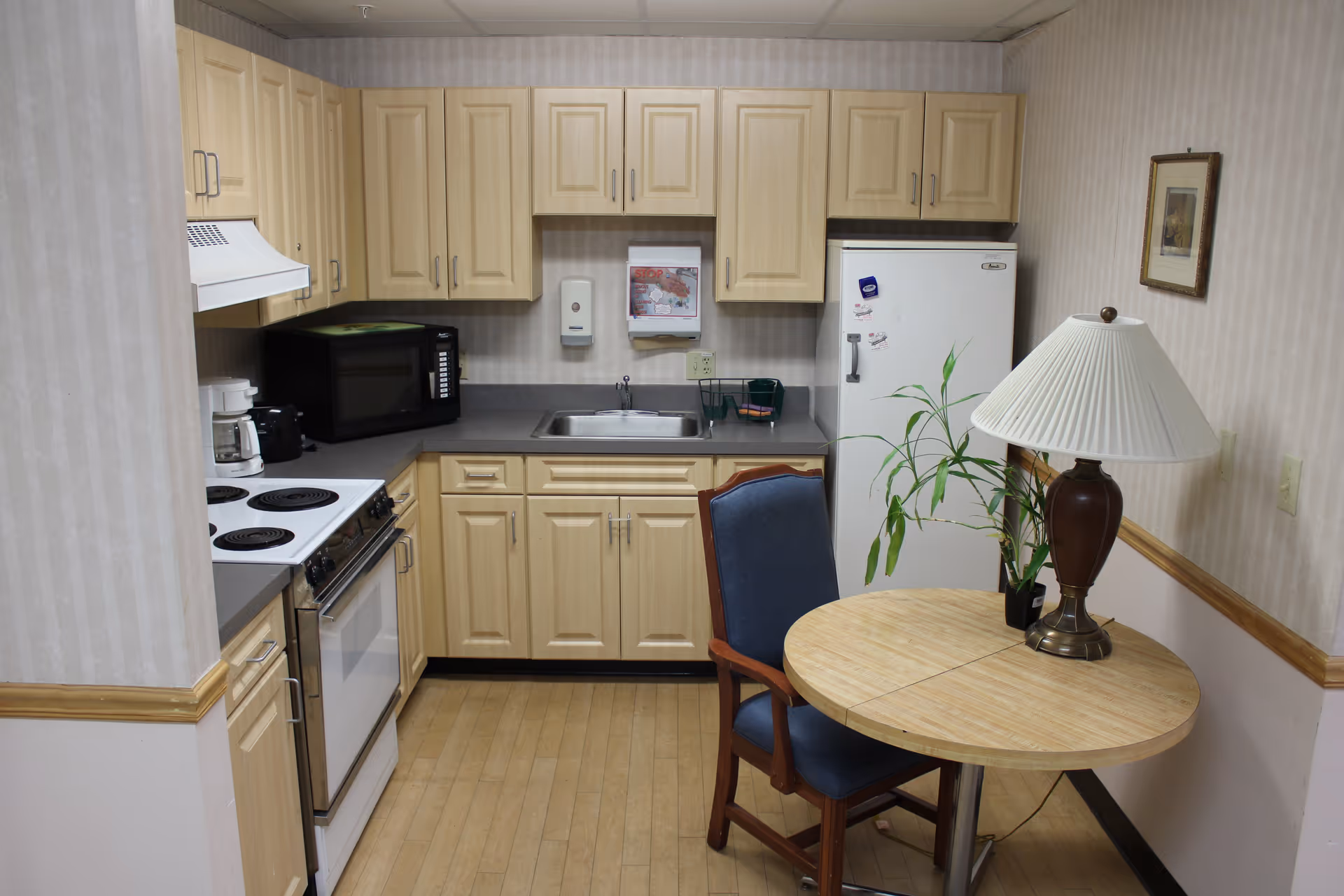A small kitchenette with light wood cabinets, a stove, microwave, sink, refrigerator, and a round table with a chair, lamp, and plant.