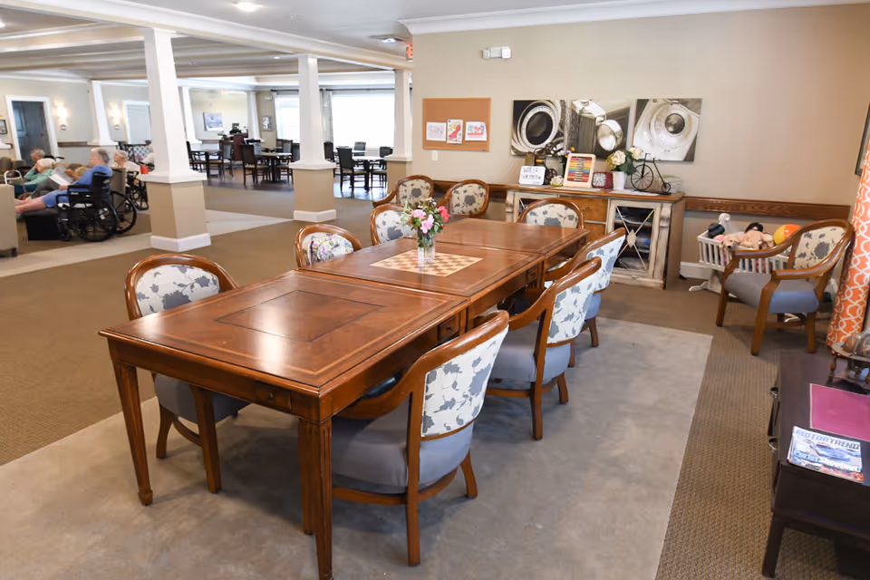 A spacious dining room with a large wooden table surrounded by upholstered chairs, floral decorations, and artwork on the walls. In the background, residents are seated in a lounge area.