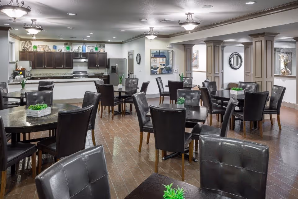 A spacious dining area with multiple dark wooden tables and black leather chairs. Each table has a small green plant centerpiece. In the background, there is a modern kitchen with dark cabinets, stainless steel appliances, and decorative items on top of the cabinets. The room has wooden flooring, white walls, and ceiling lights providing bright illumination.