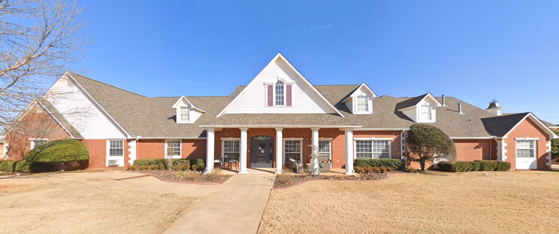 Front exterior of a single-story brick assisted living building with a central columned entrance and a paved walkway.