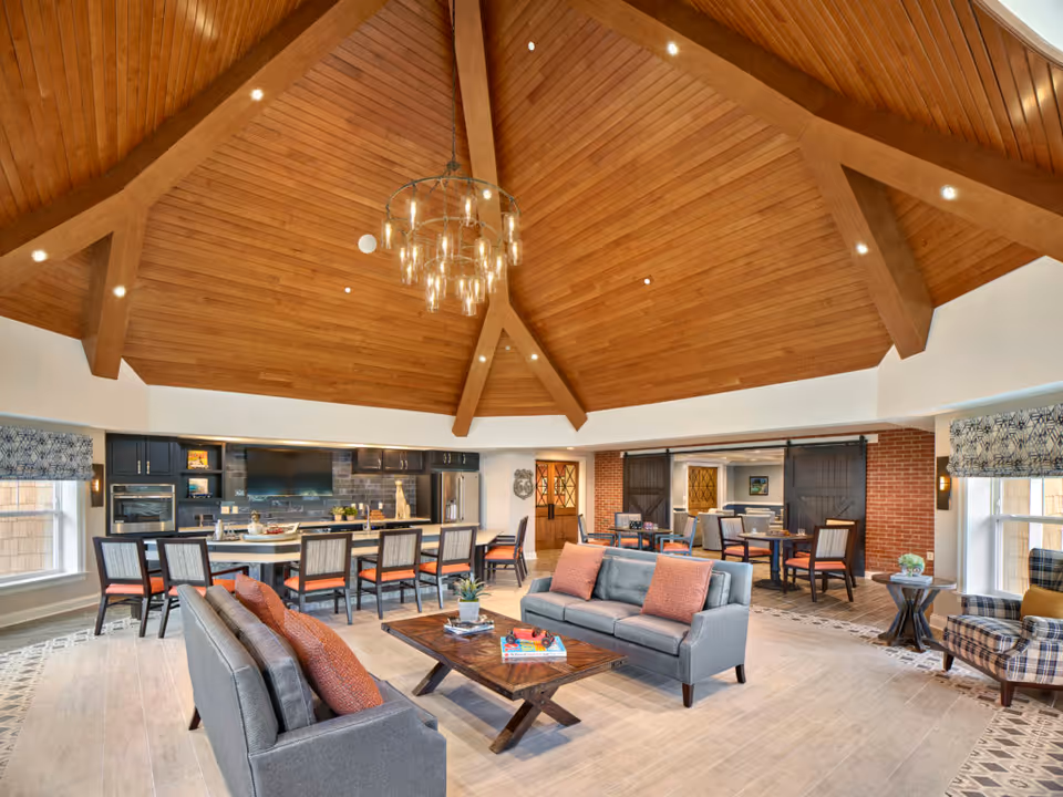 Spacious senior living common area with a high wooden vaulted ceiling and a large chandelier. The room features a kitchen area with a long island and bar stools, a seating area with two gray sofas and a wooden coffee table, and additional tables and chairs near brick walls and large windows with patterned curtains.