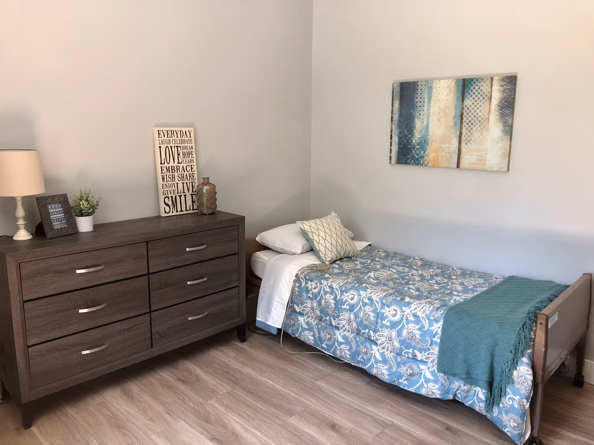 Simple furnished bedroom with a single bed, dresser, lamp, and wall art.