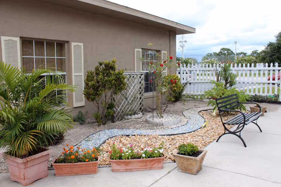Outdoor garden area next to a building with beige walls and windows with shutters. The garden features various plants and flowers in pots and planted in the ground, a small rock and gravel pathway, and a black metal bench on a concrete patio. A white picket fence and parking lot are visible in the background under a cloudy sky.