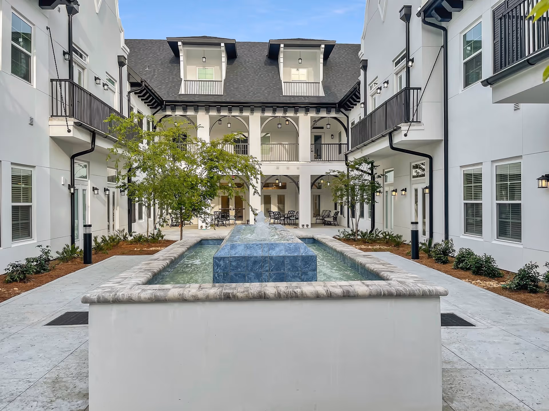 Outdoor courtyard area of The Blake at Miramar Beach featuring a central rectangular water fountain with blue tiles, surrounded by a paved walkway, small trees, and white multi-story buildings with balconies and windows.
