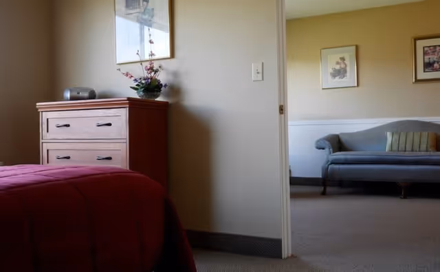 View from a bedroom showing part of a bed with a red blanket, a wooden dresser with a small radio and a flower arrangement on top, and an adjacent room with a gray upholstered sofa and framed pictures on the wall.