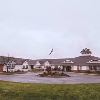 Exterior view of a single-story senior living facility building with a circular driveway and a flagpole in the center. The building has multiple peaked roof sections and a cupola on the right side. The sky is overcast and there is a grassy area in the foreground.