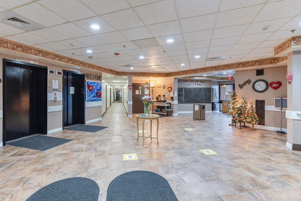 Spacious senior living facility lobby with elevators on the left, a central table with flowers, mailboxes and reception area in back, and decorated Christmas trees on the right.