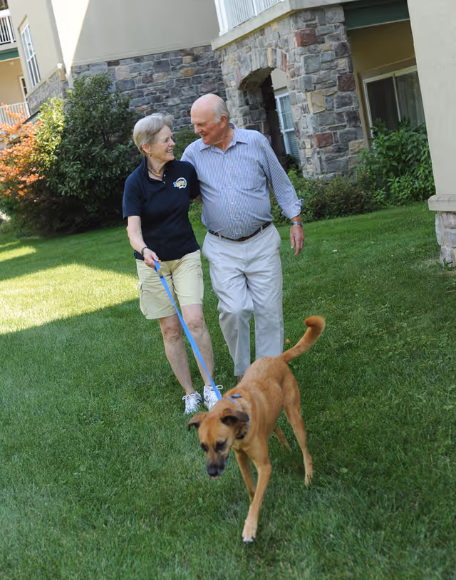 An elderly couple walking arm in arm on a grassy lawn outside a stone and stucco building, with the woman holding a leash attached to a brown dog walking ahead of them.