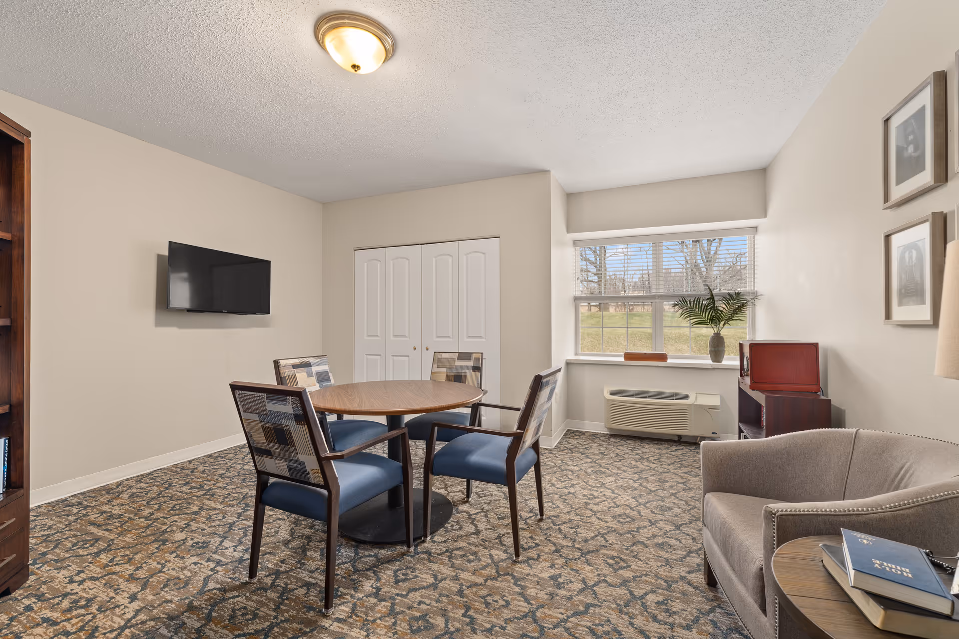 A cozy senior living room with a round wooden table surrounded by four cushioned chairs, a wall-mounted flat-screen TV, a window with a view of trees and grass, a small cabinet with a plant on top, and a comfortable armchair next to a side table with books.
