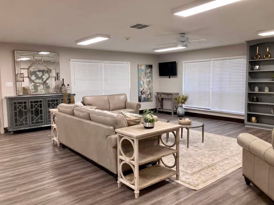 Bright communal living room with beige sofas, coffee tables, an area rug, shelving, and large windows with blinds.