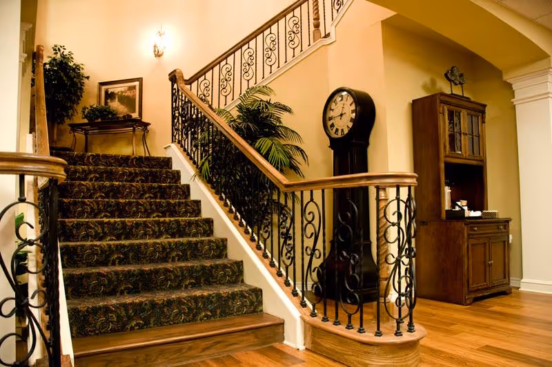 Interior view of a senior living facility featuring a carpeted staircase with ornate black iron railings and wooden handrails. To the right of the stairs is a tall grandfather clock and a wooden cabinet with glass doors. There is a potted plant near the staircase and a framed picture on the wall above a small table. The flooring is wooden, and the walls are painted a warm beige color.
