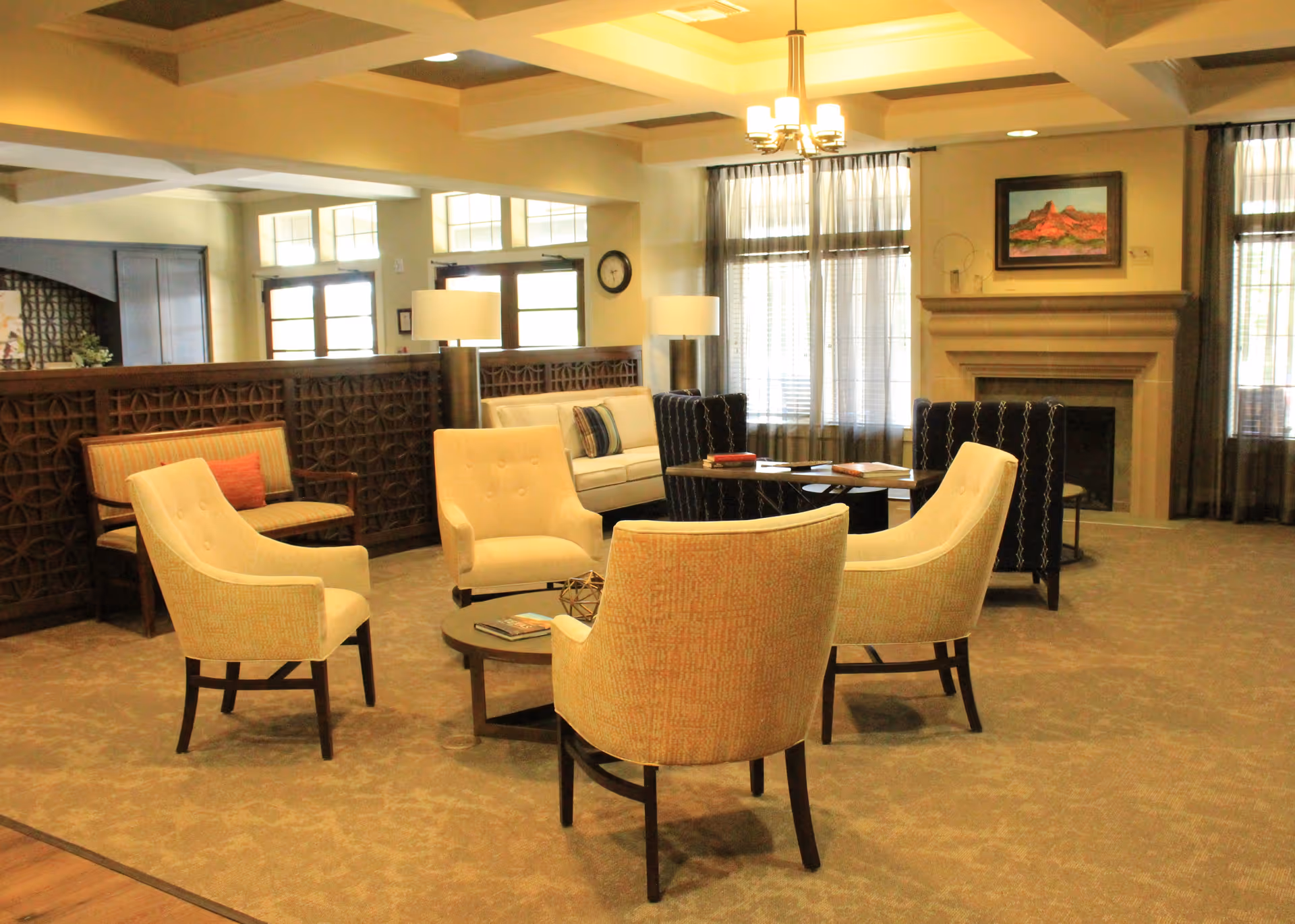 A spacious and well-lit living room area with beige carpet and a coffered ceiling. The room features a circular arrangement of four beige upholstered armchairs around a round coffee table with books and decorative items. Behind the chairs, there is a white sofa with a striped pillow, two black patterned armchairs, and two tall lamps. Large windows with sheer curtains allow natural light to fill the room. A fireplace with a framed landscape painting above it is visible on the right side of the room.