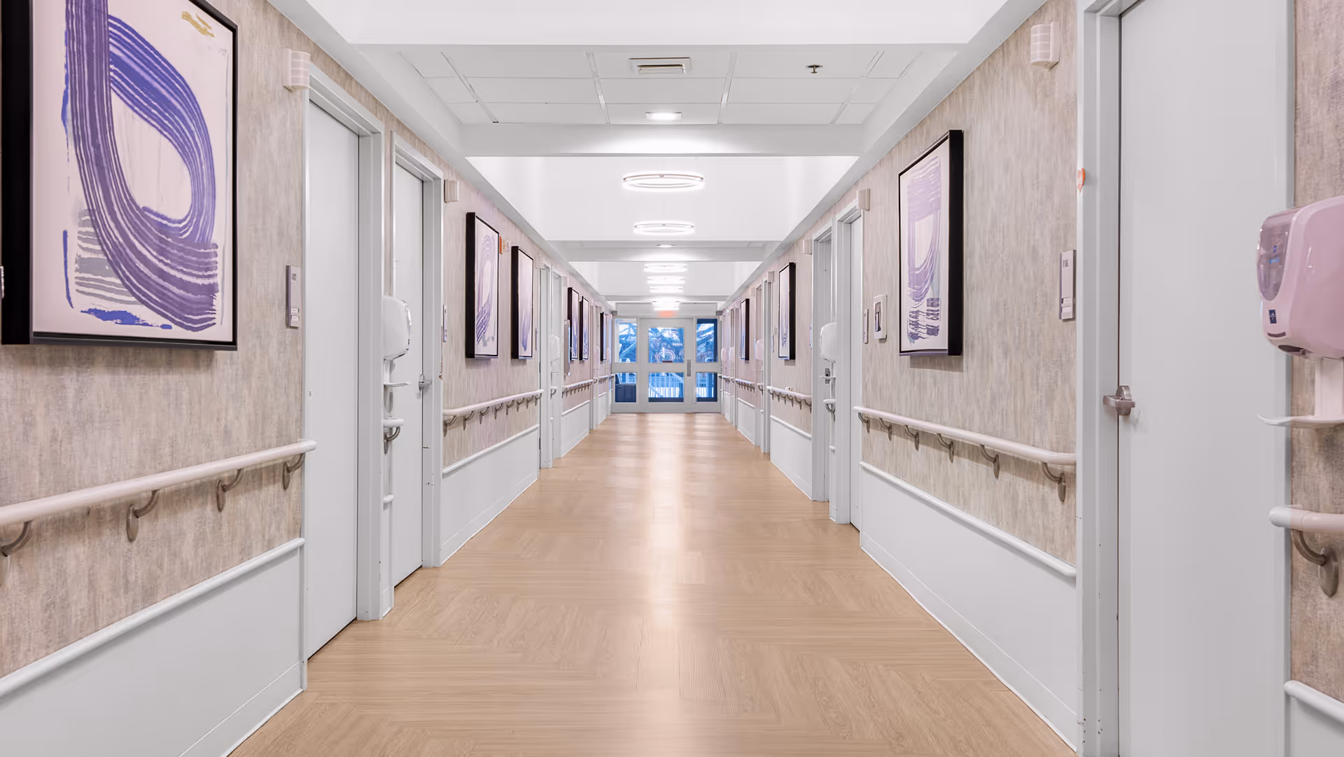 A long, well-lit hallway in a senior living facility with light wood flooring, beige walls, and handrails on both sides. Several doors line the hallway, and framed abstract purple artwork is hung on the walls. At the end of the hallway, there are large windows letting in natural light.