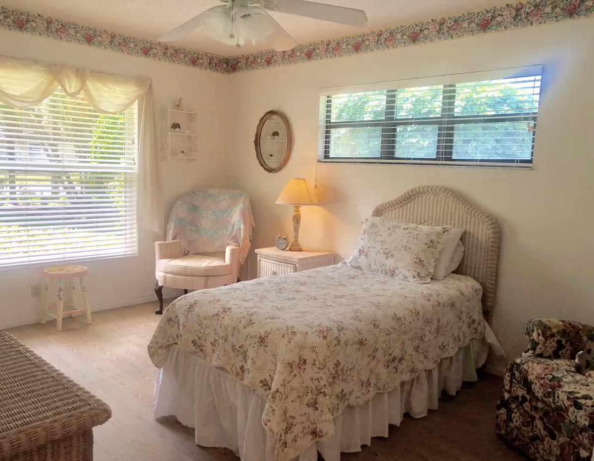 Sunlit bedroom with a single bed in floral bedding, a wicker headboard, nightstand with lamp, armchair, and large windows with blinds.