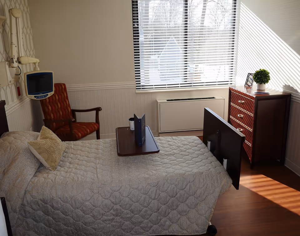 A well-lit room with a single hospital-style bed covered with a beige quilted blanket and a decorative pillow. Next to the bed is a red patterned armchair and a mounted medical monitor. A small wooden table is placed on the bed with a cup and a menu or booklet. A window with blinds lets in natural light, illuminating a wooden dresser with a small green plant and a framed photo on top.