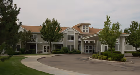 Front exterior view of Brookdale Boise Parkcenter, a two-story building with a covered entrance, surrounded by trees and landscaped greenery under a cloudy sky.