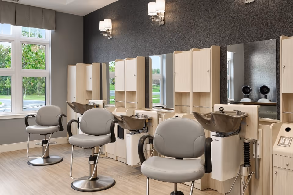 Interior view of a hair salon area with three gray salon chairs in front of wooden stations equipped with sinks and mirrors. Large windows on the left side let in natural light, and the walls are painted gray with modern light fixtures above the stations.