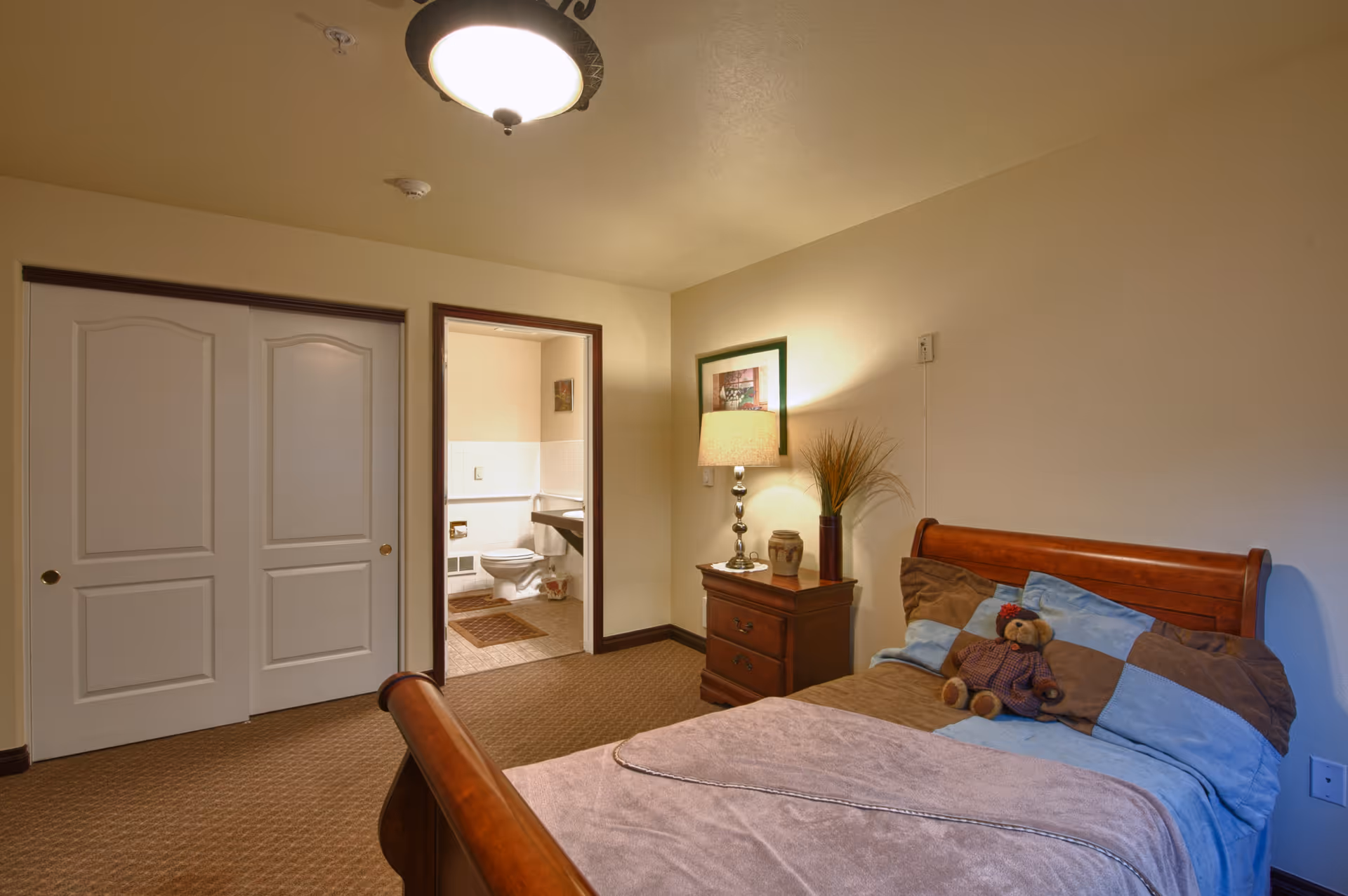 A cozy bedroom with a wooden bed featuring a patchwork quilt and a teddy bear on the pillows. Next to the bed is a wooden nightstand with a lamp, a decorative vase, and a framed picture on the wall. The room has beige walls and carpeted floor. A doorway leads to a bathroom with a toilet and a sink visible. There is also a closed closet with white double doors.