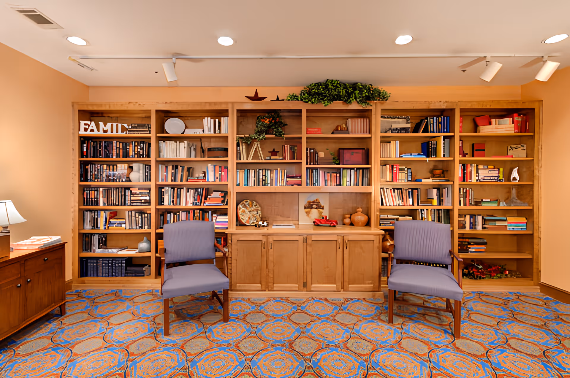 A cozy reading or sitting area with a large wooden bookshelf filled with books and decorative items. Two purple upholstered chairs are placed in front of the bookshelf on a patterned carpet with blue and orange designs. A wooden sideboard with a lamp and some books is visible on the left side of the image. The walls are painted a warm beige color and the ceiling has recessed lighting.