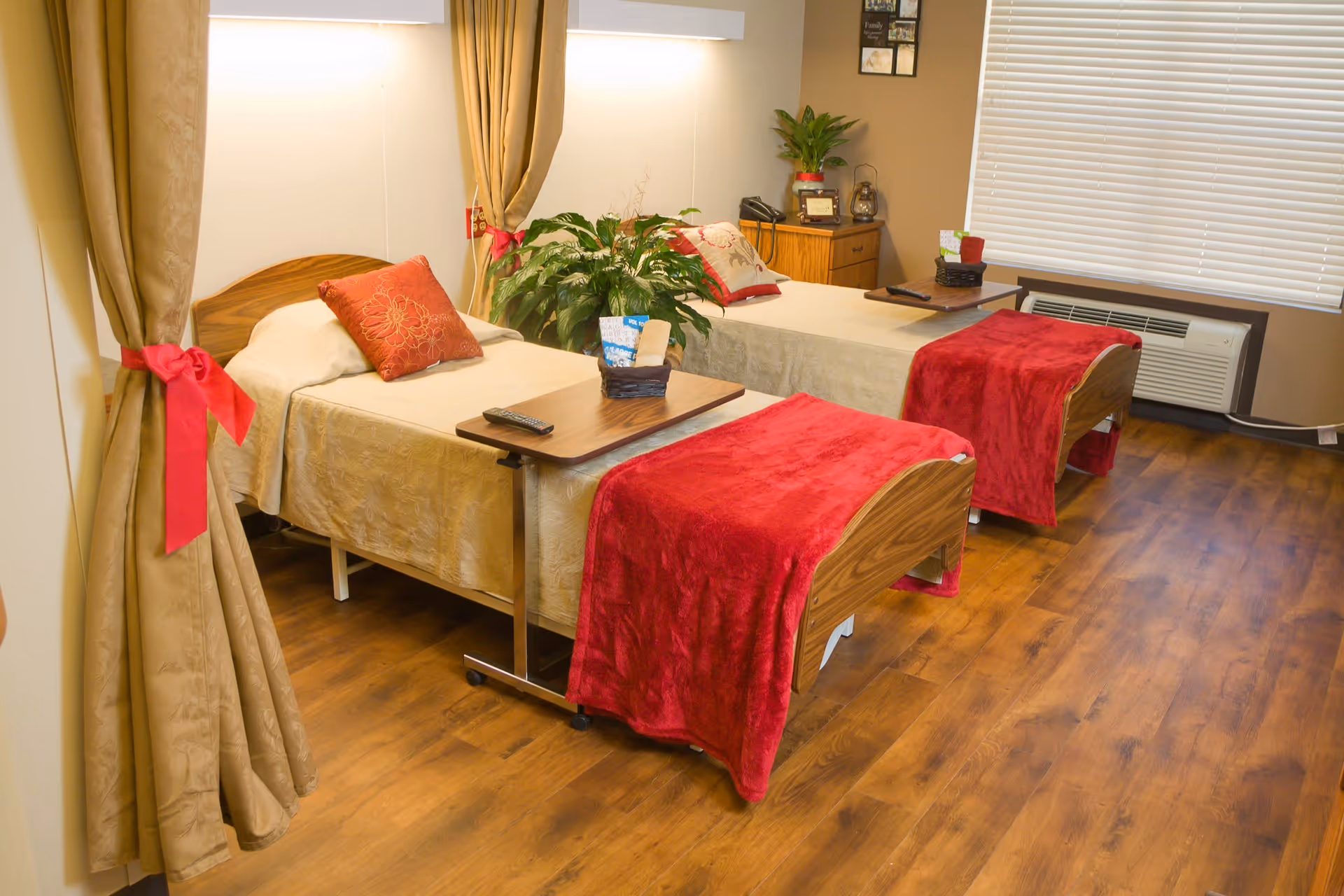 A cozy room with two single beds, each covered with beige bedspreads and red throws at the foot. Each bed has a red decorative pillow and a small overbed table with items on top. There is a large green plant between the beds, beige curtains tied back with red ribbons, wooden flooring, and a window with blinds and an air conditioning unit below it.