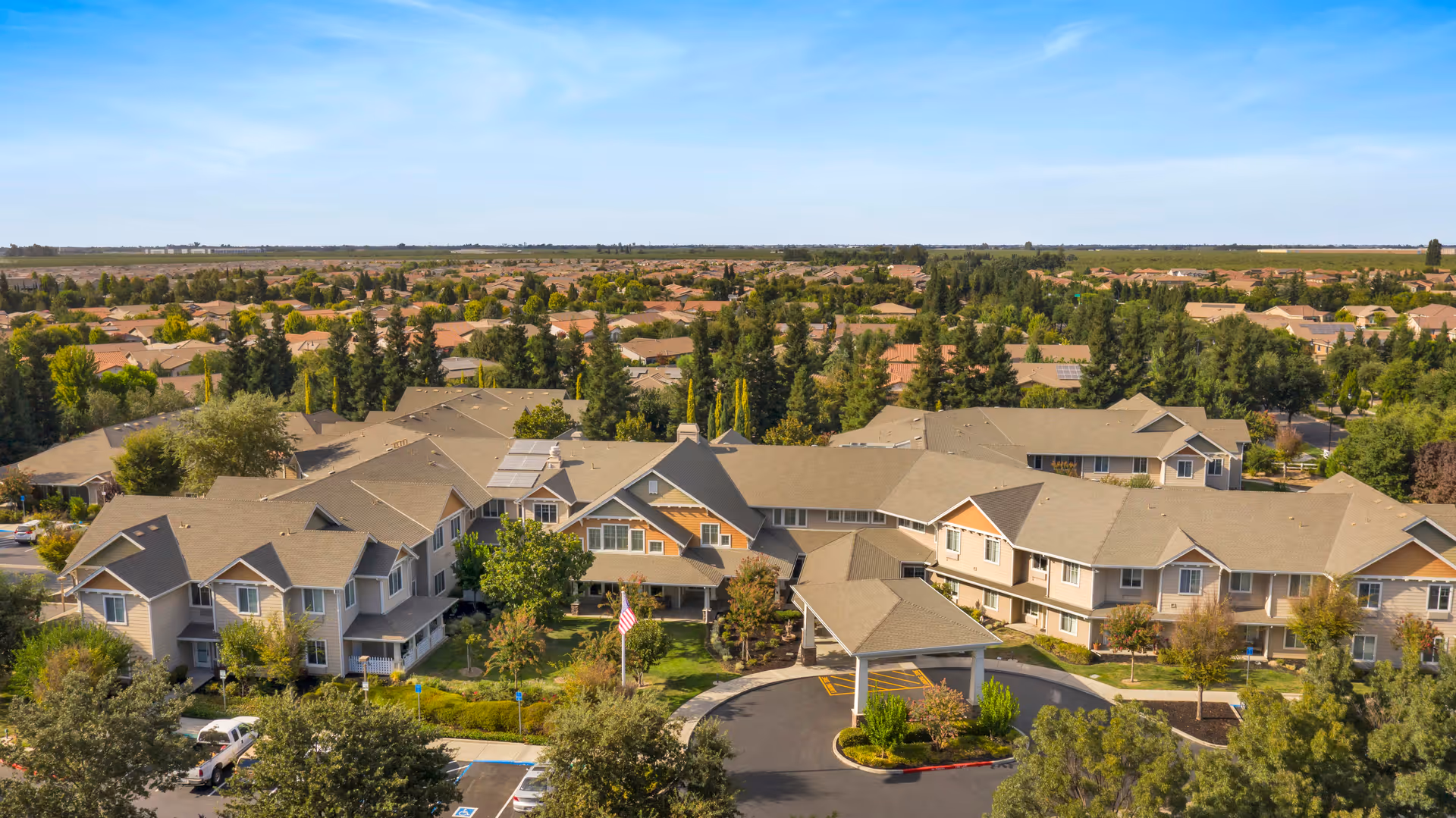 Aerial view of a senior living facility named The Commons at Union Ranch, showing multiple connected buildings with beige roofs surrounded by trees and greenery, with a parking area and an American flag in front.