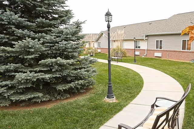Curved concrete walkway bordered by green grass and a large evergreen tree on the left. There are black lamp posts along the path and a bench with cushions on the right side. In the background, there is a single-story building with beige siding and a brick lower exterior.