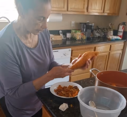 An elderly woman in a purple long-sleeve shirt is peeling a sweet potato in a kitchen. She is standing at a counter with a bowl of sweet potato peels, a large pot, and a plastic mixing bowl with a spoon inside. The kitchen has wooden cabinets, a dishwasher, and various kitchen items on the counter.