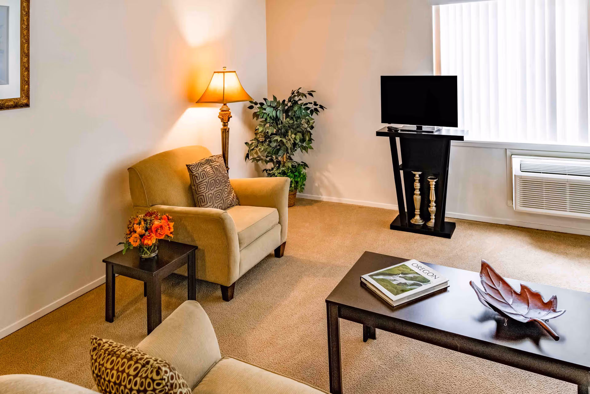 A cozy living room with beige carpet and walls. There are two beige armchairs with patterned cushions, a small dark wooden side table with a vase of orange flowers, a floor lamp, a potted plant in the corner, a flat-screen TV on a modern black stand, and a dark wooden coffee table with a book titled 'OREGON' and a decorative leaf-shaped bowl.