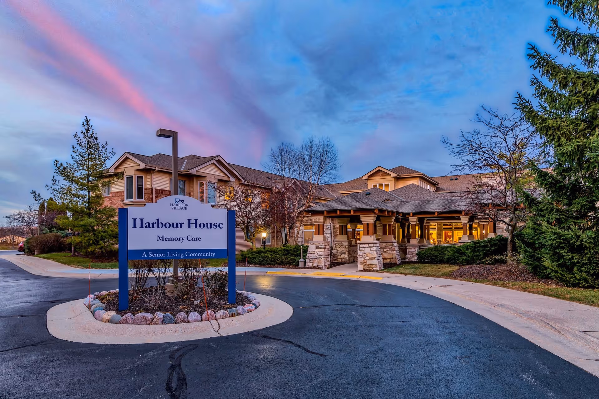 Exterior view of Harbour House Memory Care, a senior living community, during twilight with a clear sky showing pink and blue hues. The building is surrounded by trees and landscaping, with a paved driveway leading to the entrance. A sign in front reads 'Harbour House Memory Care A Senior Living Community.'