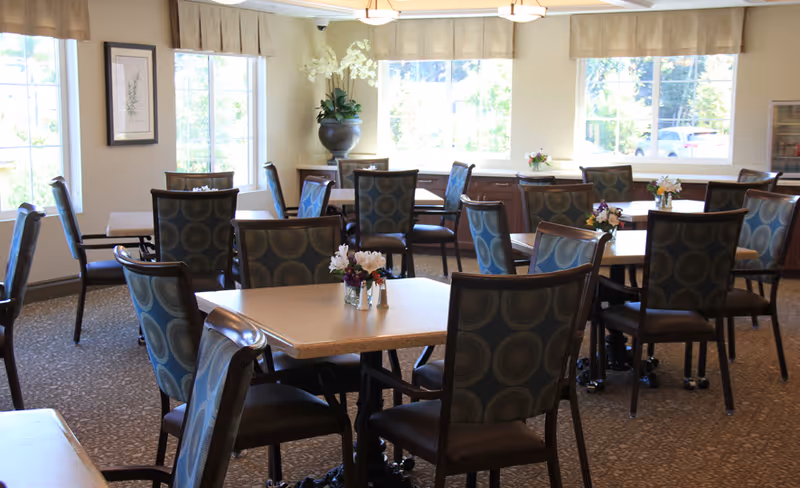 A bright dining room with multiple square tables and cushioned chairs arranged neatly. Each table has a small floral centerpiece. Large windows with beige valances allow natural light to fill the room. There is a large decorative vase with white flowers in the corner and framed artwork on the wall.