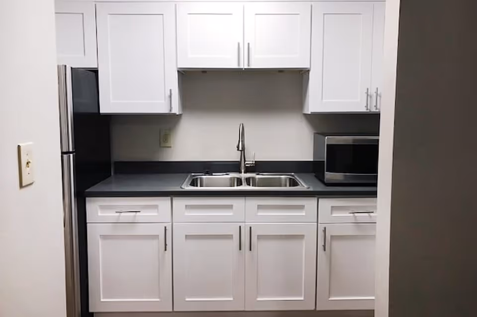 A small kitchen area with white cabinets and drawers, a double stainless steel sink with a modern faucet, a microwave on the right countertop, and a stainless steel refrigerator on the left side.