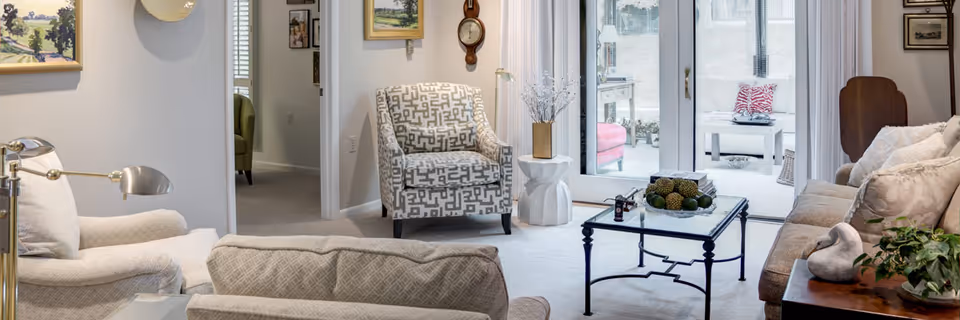 A cozy living room with beige and patterned armchairs, a glass coffee table with decorative items, and a sofa with cushions. The room has large glass doors leading to a bright outdoor patio area with seating. The walls are decorated with paintings and a clock.