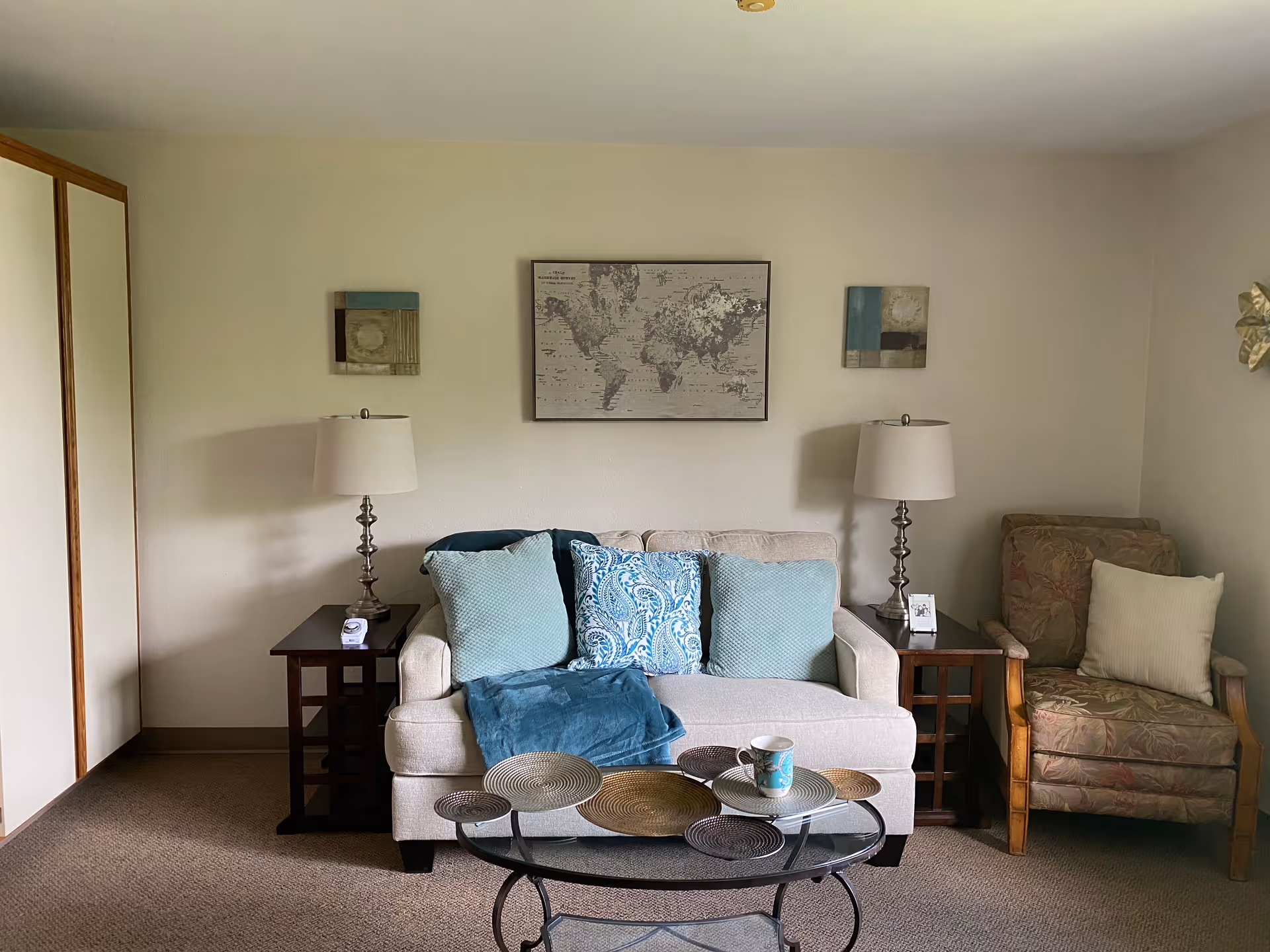 A cozy living room with a beige loveseat adorned with three blue and patterned throw pillows and a blue throw blanket. On either side of the loveseat are dark wooden side tables with matching lamps. To the right is a floral-patterned armchair with a white pillow. A glass coffee table with decorative round placemats and a mug sits in front of the loveseat. The walls are light-colored and decorated with a world map and two small abstract paintings.